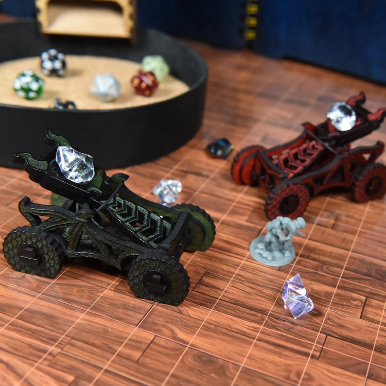 May include: Two miniature wooden catapults, one dark green and one dark red, each featuring a clear crystal projectile. A small gray figurine stands nearby on a brown grid surface, with a circular tray containing several multi-colored dice.