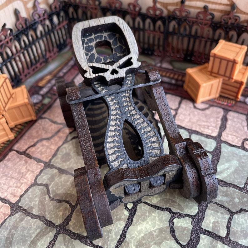 May include: A wooden toy catapult with a skull design. The catapult is made of dark brown wood and has a black and white skull design on the front. The catapult is in focus and the background is blurred.