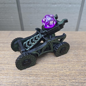 May include: A black and dark green 3D-printed catapult with a dragon-inspired frame and four textured wheels. A translucent purple twenty-sided die with white numbers is positioned in the cradle of the catapult, ready to be launched.