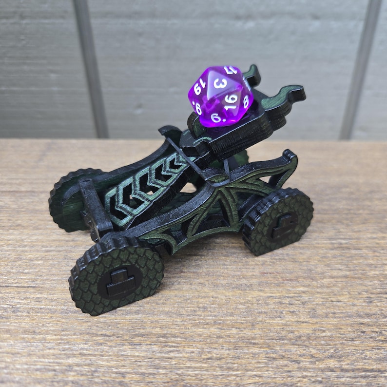 May include: A black and dark green 3D-printed catapult with a dragon-inspired frame and four textured wheels. A translucent purple twenty-sided die with white numbers is positioned in the cradle of the catapult, ready to be launched.