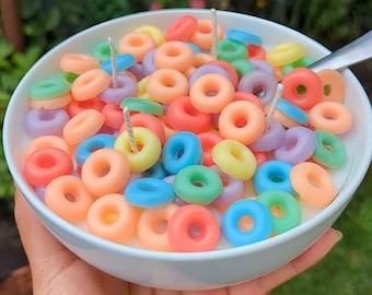 May include: A white ceramic bowl filled with a white wax base and numerous small, colorful, ring-shaped wax pieces in shades of pink, orange, yellow, green, blue, and purple. Three white candle wicks protrude from the wax cereal mixture.