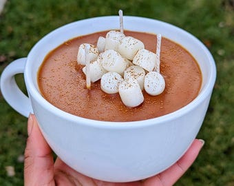 Puede incluir: Una taza de cerámica blanca llena de cera marrón que parece chocolate caliente, cubierta con varias piezas pequeñas de cera blanca con forma de malvavisco y una ligera capa de polvo marrón. Tres mechas blancas finas sobresalen de la cera.