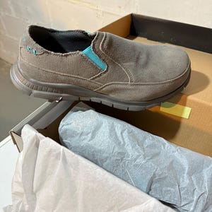 May include: A taupe slip-on shoe with a textured sole and teal accents. The shoe is in an open cardboard box with tissue paper. The shoe has a canvas-like upper and a cushioned insole.
