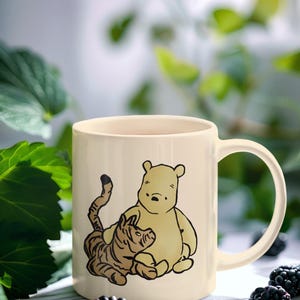 May include: A cream-colored ceramic mug with a Winnie the Pooh and Tigger illustration. The mug has a black handle and is sitting on a white surface with blackberries.
