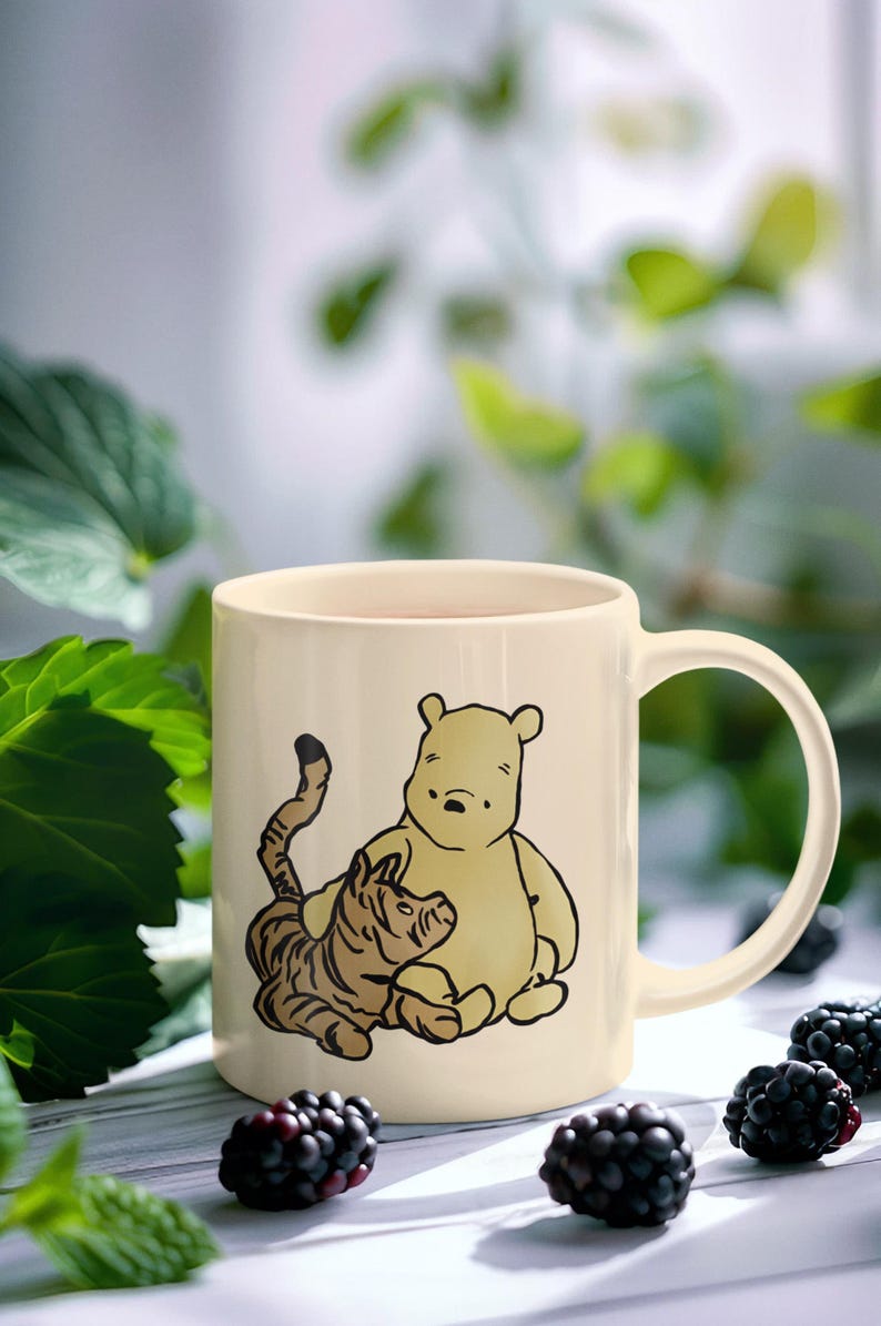 May include: A cream-colored ceramic mug with a Winnie the Pooh and Tigger illustration. The mug has a black handle and is sitting on a white surface with blackberries.