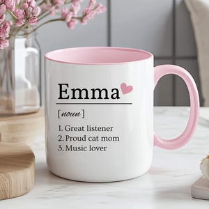 May include: White ceramic mug with a pink interior and handle. The mug features the name "Emma" with a heart graphic, followed by a definition: "Great listener, Proud cat mom, Music lover."