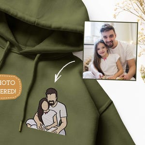 May include: Olive green hoodie with a drawstring and hood. The hoodie features an embroidered image of a couple and the text "YOUR PHOTO EMBROIDERED!". A framed photo of a man and a girl is in the background.
