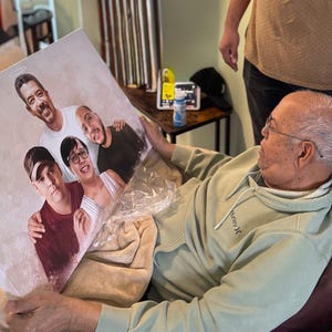 May include: A canvas print featuring a portrait of five people. The artwork depicts a group of individuals, with a halo above one person's head. The print is held by a person wearing a green hoodie.