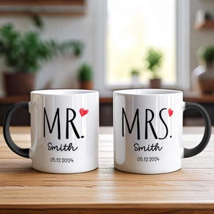 Puede incluir: Dos tazas de cerámica blanca con asas negras. Las tazas tienen las palabras "MR." y "MRS." con un corazón rojo y el texto "Smith 05.12.2024".