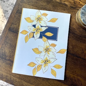 May include: A white journal with a yellow border and a floral design featuring white flowers with yellow centers and yellow leaves. A navy blue rectangle with a white border is in the upper right corner of the journal.