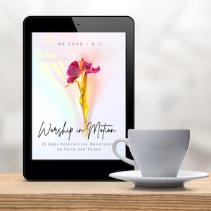 May include: A tablet computer displaying the cover of a book titled "Worship in Motion: 21 Days Interactive Devotions on Faith and Dance". The cover features a pink flower with a yellow stem against a colorful background. A white coffee cup and saucer are on a wooden table in front of the tablet.