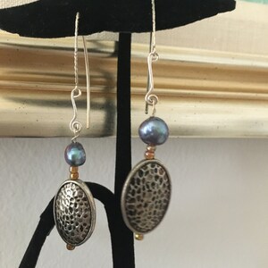 May include: A pair of silver earrings with a hammered oval charm and a dark blue pearl bead. The earrings are hanging from a silver hook.