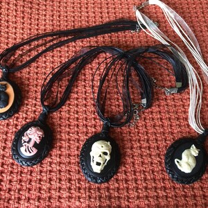 May include: Four black cameo pendants with different designs, including a cat, a skull, a woman, and a cat. The pendants are hanging from black ribbon necklaces.
