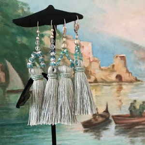 May include: Three pairs of light blue tassel earrings with crystal beads. The earrings are hanging from a black stand against a painted background of a seascape.