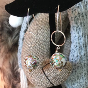 May include: A pair of silver dangle earrings with a white round bead featuring a gold and green floral design.