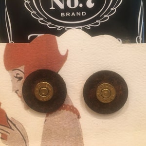 May include: A pair of brown leather earrings with a gold bullet charm. The earrings are displayed on a white card with a red-haired woman illustration.