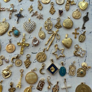 Puede incluir: Una colecci&oacute;n de dijes y colgantes dorados, que incluyen corazones, cruces, estrellas y figuras religiosas. Los dijes est&aacute;n dispuestos sobre una superficie blanca con una nota escrita a mano.