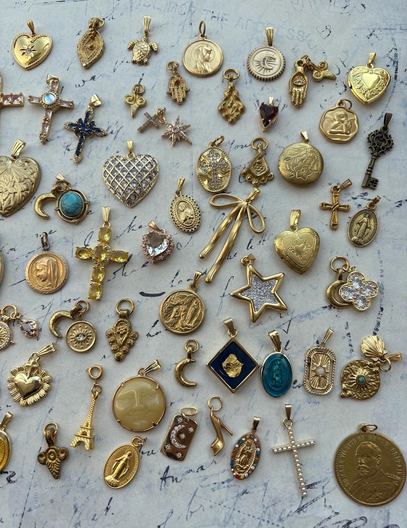Puede incluir: Una colecci&oacute;n de dijes y colgantes dorados, que incluyen corazones, cruces, estrellas y figuras religiosas. Los dijes est&aacute;n dispuestos sobre una superficie blanca con una nota escrita a mano.