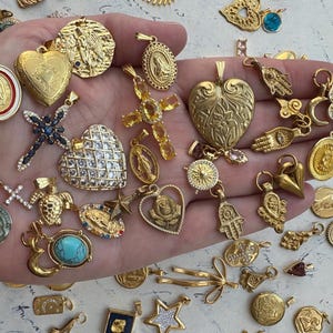 Puede incluir: Una colecci&oacute;n de dijes dorados con s&iacute;mbolos religiosos y espirituales, incluyendo cruces, corazones, manos y tortugas. Los dijes est&aacute;n adornados con cristales y piedras preciosas brillantes.