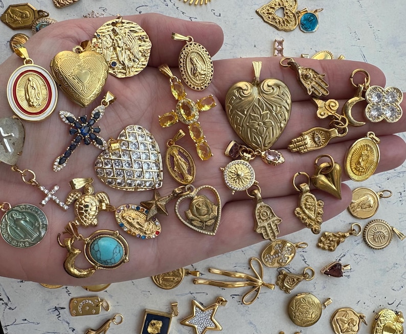Puede incluir: Una colecci&oacute;n de dijes dorados con s&iacute;mbolos religiosos y espirituales, incluyendo cruces, corazones, manos y tortugas. Los dijes est&aacute;n adornados con cristales y piedras preciosas brillantes.