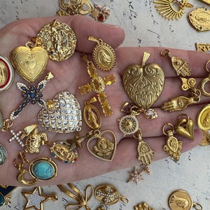 Puede incluir: Una mano sosteniendo una variedad de dijes y colgantes dorados. Los dijes incluyen corazones, cruces, estrellas, manos y otros s&iacute;mbolos religiosos. Los dijes est&aacute;n todos intrincadamente detallados y tienen una est&eacute;tica vintage.