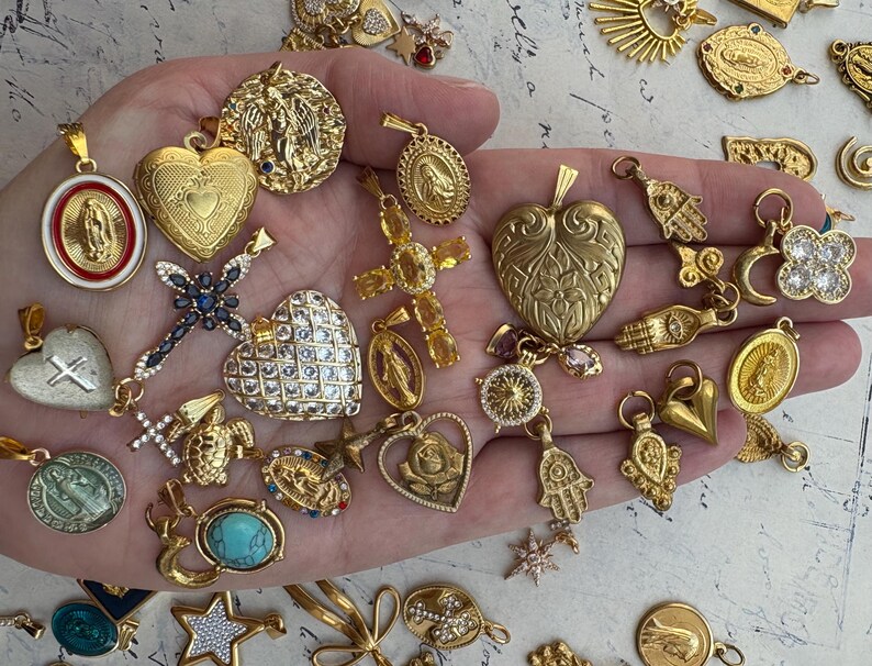 Puede incluir: Una mano sosteniendo una variedad de dijes y colgantes dorados. Los dijes incluyen corazones, cruces, estrellas, manos y otros s&iacute;mbolos religiosos. Los dijes est&aacute;n todos intrincadamente detallados y tienen una est&eacute;tica vintage.