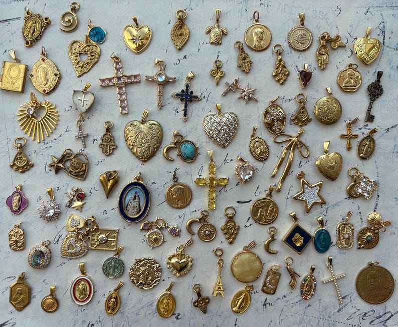 Puede incluir: Una colecci&oacute;n de dijes dorados con varios dise&ntilde;os religiosos, de la naturaleza y abstractos. Los dijes est&aacute;n dispuestos sobre una superficie de color claro.