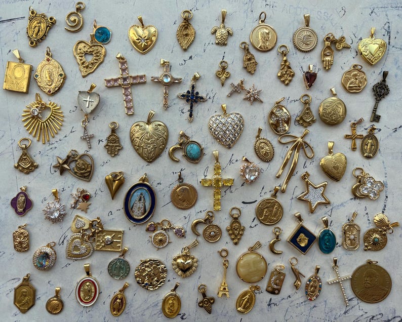 Puede incluir: Una colecci&oacute;n de dijes dorados con varios dise&ntilde;os religiosos y simb&oacute;licos, incluyendo corazones, cruces, estrellas y llaves. Los dijes est&aacute;n dispuestos sobre un fondo blanco.