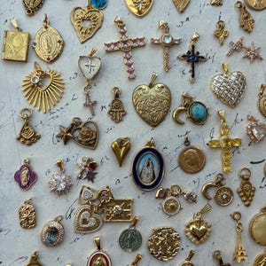 Puede incluir: Una colecci&oacute;n de dijes y colgantes dorados, que incluyen corazones, cruces, estrellas y figuras religiosas. Los dijes est&aacute;n dispuestos sobre una superficie blanca con una nota escrita a mano en el fondo.