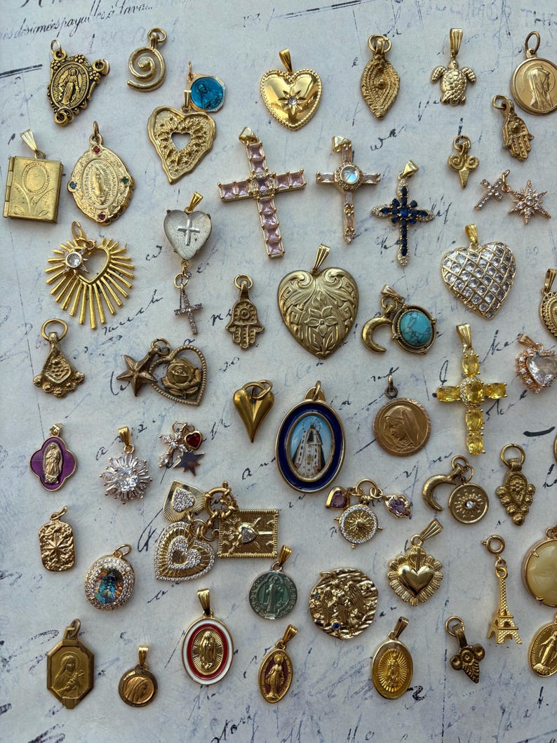 Puede incluir: Una colecci&oacute;n de dijes y colgantes dorados, que incluyen corazones, cruces, estrellas y figuras religiosas. Los dijes est&aacute;n dispuestos sobre una superficie blanca con una nota escrita a mano en el fondo.