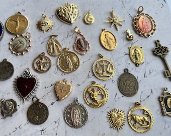 Pode incluir: Uma coleção de pingentes e amuletos religiosos dourados, incluindo um coração, uma balança da justiça, uma cruz, um leão e uma variedade de figuras religiosas. Os pingentes estão dispostos em um fundo branco com texto escrito à mão.