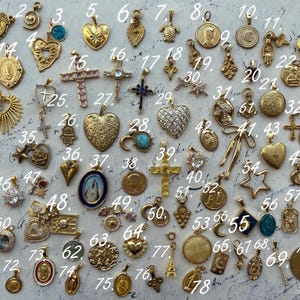 Puede incluir: Una colecci&oacute;n de 78 dijes dorados con varios dise&ntilde;os religiosos y simb&oacute;licos. Los dijes est&aacute;n dispuestos en filas sobre una superficie blanca. Algunos de los dijes presentan corazones, cruces, estrellas y otros s&iacute;mbolos religiosos.