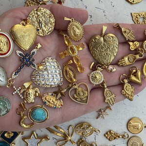Puede incluir: Una colecci&oacute;n de dijes dorados con s&iacute;mbolos religiosos y espirituales, incluyendo corazones, cruces, manos y una variedad de otros dise&ntilde;os. Los dijes se muestran en una mano.