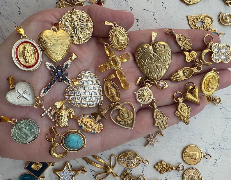 Puede incluir: Una colecci&oacute;n de dijes dorados con s&iacute;mbolos religiosos y espirituales, incluyendo corazones, cruces, manos y una variedad de otros dise&ntilde;os. Los dijes se muestran en una mano.