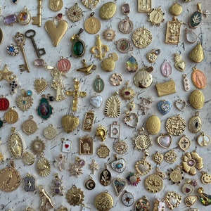 Op de afbeelding: Een verzameling goudkleurige hangers en bedels, waaronder kruisen, harten, sleutels en religieuze symbolen. Sommige hangers hebben kleurrijke edelstenen en ingewikkelde ontwerpen. De items zijn gerangschikt op een getextureerd, gebroken wit oppervlak.