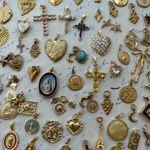 Puede incluir: Una colecci&oacute;n de dijes dorados con diversos dise&ntilde;os religiosos y simb&oacute;licos, incluyendo corazones, cruces, estrellas y una luna creciente. Los dijes est&aacute;n dispuestos sobre una superficie blanca con texto escrito a mano en el fondo.