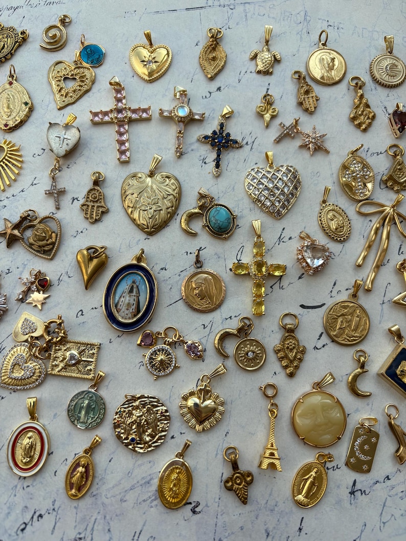 Puede incluir: Una colecci&oacute;n de dijes dorados con diversos dise&ntilde;os religiosos y simb&oacute;licos, incluyendo corazones, cruces, estrellas y una luna creciente. Los dijes est&aacute;n dispuestos sobre una superficie blanca con texto escrito a mano en el fondo.