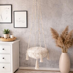 Könnte beinhalten: Ein hängendes Katzenbett aus Makramee mit einem flauschigen, weißen Kissen. Das Bett wird von der Decke abgehängt und hat dekorative Quasten. Der Raum ist in neutralen Farben gehalten und verfügt über eine weiße Kommode, Katzenkunst und eine Vase mit Trockenblumen.