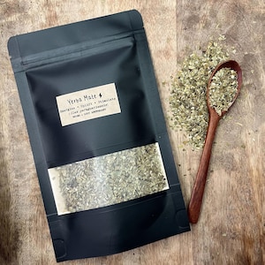 May include: A black pouch of Yerba Mate tea with a clear window, a wooden spoon, and a pile of loose tea leaves. The label reads "Yerba Mate" with the words "Energize, Uplift, Stimulate". The background is a wooden surface.
