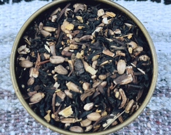 May include: A round, metallic gold-toned tin filled with a loose-leaf tea blend. The mixture consists of dark, dried tea leaves, light tan seeds, and small, woody, fibrous pieces, creating a textured appearance within the circular container.