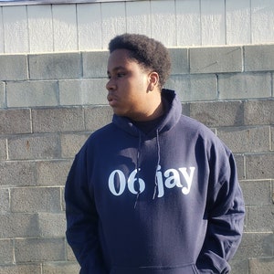 Puede incluir: Una sudadera azul marino con un gráfico blanco que dice "06 Jay".