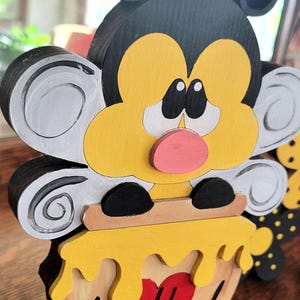 May include: A wooden bee-themed decoration with a yellow face, black antennae, and white and gray wings. The bee sits atop a honey pot with the words "bee kind" and a red heart. The design is painted with bright colors.