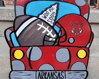 May include: Red pickup truck with white polka dots, a football, and a red helmet with a Razorback logo. The truck has a blue bed and the words "Woo Pig Sooie" and "Arkansas" are written on the truck.