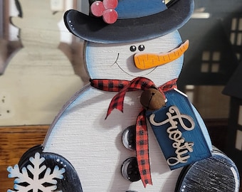 May include: A wooden snowman decoration featuring a black top hat with a white snowflake, red berries, and green pine needles. The snowman wears a red and black buffalo plaid scarf with a small bell and a blue tag labeled Frosty.