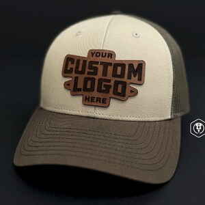 May include: A beige and brown trucker hat with a brown leather patch that says "YOUR CUSTOM LOGO HERE" in a bold font.