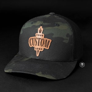 May include: A black and green camouflage trucker hat with a brown leather patch that says "YOUR CUSTOM LOGO" in a vintage font.