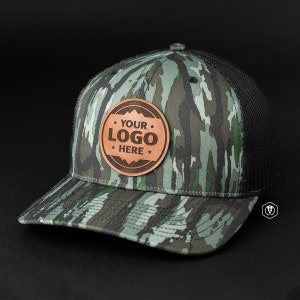 May include: A camouflage trucker hat with a black mesh back. The hat has a brown leather patch with the text "YOUR LOGO HERE" in white.