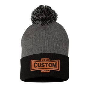 May include: Gray and black knit beanie with a black pom pom on top. The beanie has a brown leather patch with the text "YOUR CUSTOM LOGO" on the front.