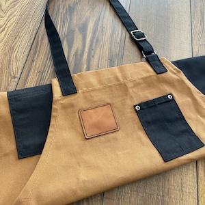 May include: A brown canvas apron with black straps and a black pocket. The apron has a brown leather patch on the front.