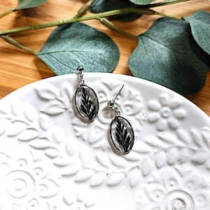 May include: Silver-toned dangle earrings with oval-shaped clear resin pendants. Each pendant contains a black fern leaf. The earrings are displayed on a white, floral-patterned plate, with green leaves in the background.